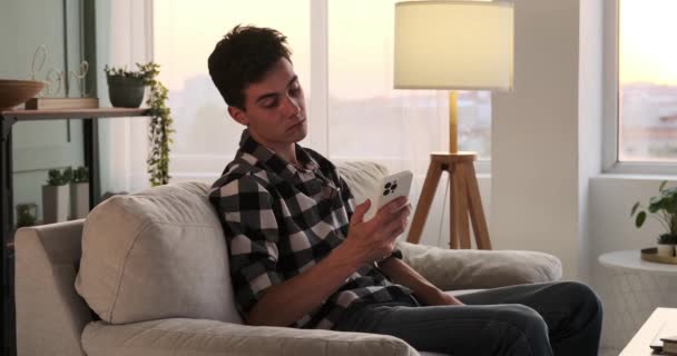 在客厅里 一个白种人躺在沙发上 一边翻看手机 一边显得很无聊 空气是一种悠闲的气氛 他的举止带有一丝不安的意味 — 图库视频影像