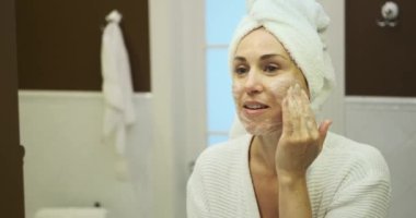 Aynanın karşısında duran bir kadın, ustalıkla yüz temizleyicisini uyguluyor ve bunu bir amaç için cildine uyguluyor. Bu kişisel bakım anı, sağlıklı bir cilt bakımına olan bağlılığını gösteriyor..