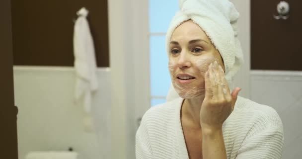 站在镜子前 一个女人巧妙地使用面部清洁剂 有目的地把它涂在皮肤上 这个自我保健的时刻展现了她对保持肌肤健康日常护理的承诺 — 图库视频影像