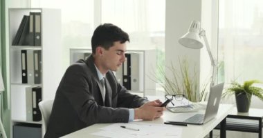 Neşeli iş adamı masasında otururken telefonunu kullanırken görülüyor. Parlak bir gülümseme ve olumlu bir tavırla cihaza neşeli bir şekilde bağlanır..