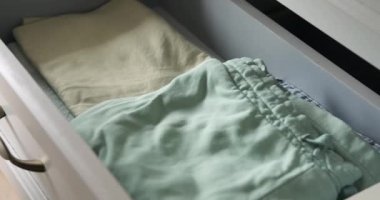 Bir çift kadın eli, elbiseleri titizlikle katlar ve onları şifoniyer çekmecesine nazikçe yerleştirir. Düzenli hareketler, organizasyona bilinçli bir yaklaşımdan bahseder..