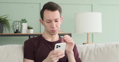 Bu şaşkın anda, oturma odasındaki kanepede oturan bir adam resmediliyor, telefonunu kullanırken şaşkın bakıyor. Yüz ifadesi kafa karışıklığını ve belirsizliği yansıtıyor..