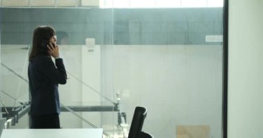 Pencerenin yanında bir telefon görüşmesi yaparken sakin ve odaklanmış bir iş kadını. Ciddi bir ifade ve özenli bir duruşla profesyonel ve etkili bir şekilde iletişim kuruyor..