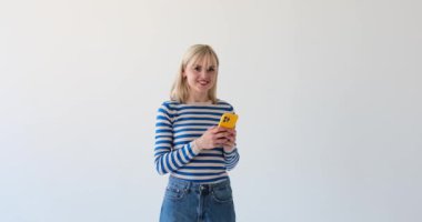 Kafkasyalı sarışın bir kadın temiz beyaz bir arka planda, telefonunu tutarken, doğrudan kameraya bakarken ve sıcak bir şekilde gülümserken görülüyor. Onun doğrudan bakışı izleyiciyle bir bağlantı hissi yaratıyor..