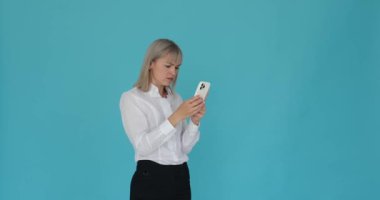 Kafası karışmış bir iş kadını telefonunu mavi arka planda kullanırken üzgün hissediyor. Çatlak kaşları ve sorunlu ifadesi kafa karışıklığını gösteriyor..