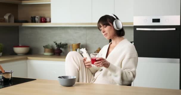 一个女人 带着她的电话和耳机 花点时间在厨房里品尝一杯饮料 她平静的表情反映了在舒适的环境中喝一杯的简单快乐 — 图库视频影像