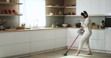 Tasasız, beyaz bir kadın, kulaklık takıyor, ev işleriyle eğlenceyi birleştiriyor mutfakta yerleri süpürürken ve dans ederken. Neşeli ve kontrolsüz hareketleri eğlenceli bir atmosfer yaratıyor..