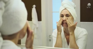 Beyaz bir kadın aynanın önünde temizleyici köpük kullanarak yüzünü nazikçe yıkar. Özenli bakımı, taze ve yenilenmiş bir cilt için kendini adamış bir cilt bakımını yansıtıyor..