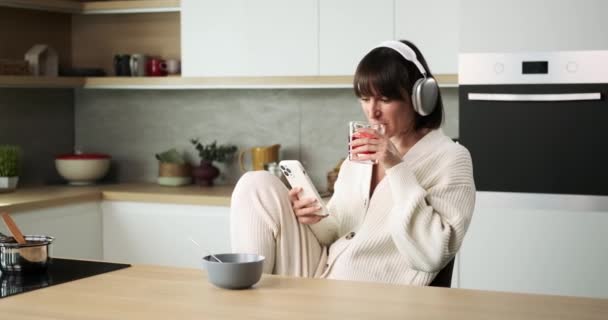 一个快乐的女人一边在厨房里一边喝着饮料一边愉快地使用她的电话 她快乐的举止和清爽的饮料使她度过了一个轻松愉快的时刻 — 图库视频影像