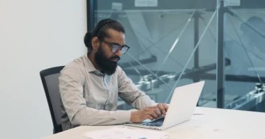 Ofisin profesyonel ortamında, Hintli bir işadamı dizüstü bilgisayarına odaklanıyor. Amaçlı tuş darbeleriyle dijital peyzajlarda geziniyor..