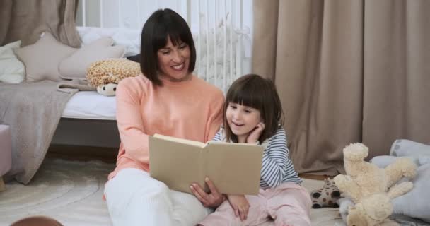 在迷人的儿童房里 一个母亲和一个女儿坐在地板上 完全沉浸在一起看书的喜悦之中 他们共同的文学冒险创造了一幅美丽的风景 — 图库视频影像