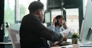 Siyahi bir işadamı, ofiste işbirliği yapan meslektaşlarına belgeleri verirken görülüyor. Bu sahne, onların etkili takım çalışmalarını ve iletişimlerini profesyonel bir ortamda yansıtıyor..