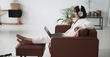 Sandalyeye oturmuş bir kadın video görüşmesi sırasında dostça el sallayarak birini selamlıyor. Onun samimi jesti ve sıcak gülümsemesi sanal buluşmada bir bağ ve dostluk duygusu iletir..