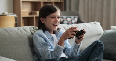 Oyun alanı ile donatılmış neşeli beyaz çocuk, kanepede otururken video oyununa dalıyor. Mutluluğu ve oyun hünerleriyle eğlenceli ve yetenekli bir oyun ortamı yaratıyor..