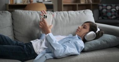 Okul çağındaki çocuk koltuğa uzanmış, elinde telefonla uzuvlarını gevşek bir ifadeyle esnetiyor. Bu boş zamanlarında, rahatlık ve rahatlık buluyor, teknoloji ve fiziksel rahatlığı birleştiriyor..