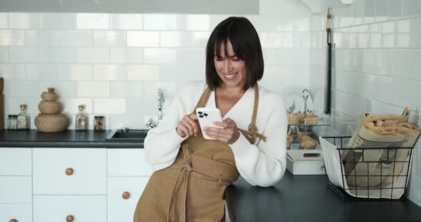 陽気な白人女性は 携帯電話を使用してエプロンに寄付され キッチンで心のこもった笑いを共有します この楽しいシーンは料理の情熱と現代のコミュニケーションのブレンドを体現しています — ストック動画