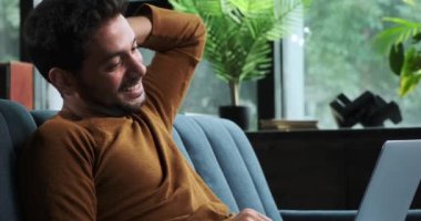 Orta Doğulu bir adam bir kanepede oturuyor, dizüstü bilgisayarını kullanıyor ve bir anlık kahkaha atıyor. Rahat ve neşeli tavrı, rahat bir ev ortamındaki iş ve eğlence dengesini temsil ediyor..