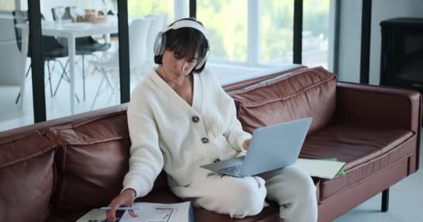 コーカサス人の女性はソファーに座り ラップトップや新聞に囲まれた仕事に囲まれています 彼女の勤勉で焦点を当てた Demeanor タスクへのコミットメントを強調し 生産性のイメージを作成する — ストック動画