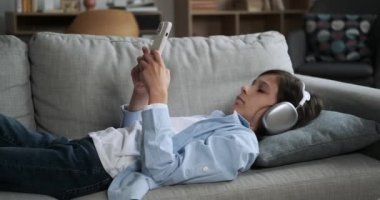 Sıkıcı beyaz çocuk, elinde telefonla kanepede uzanıyor, yorgun bir şekilde esniyor. İlgisizliği ortada, huzursuzluk ve sıkıntı anı yaratıyor..