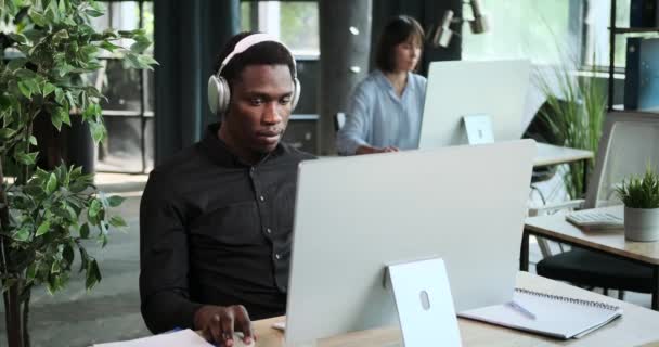 ヘッドフォンを着ている黒人ビジネスマンは 仕事を続けるにつれて音楽を楽しんでいます このシーンは 仕事中の音楽を通してモチベーションを見つけ 集中する能力を強調しています — ストック動画