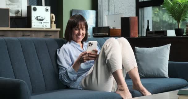 陽気な白人女性は スマートフォンを持って よく任命されたリビングルームのソファーに快適に座っています 彼女の明るい笑顔は モバイルデバイスと関わるように コンテンツとリラクゼーションを反映しています — ストック動画