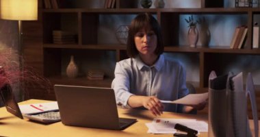 Kadın, evde oturup ödevlerini özenle yapıyor. Oda hafifçe aydınlatılıyor ve odaklanmış bir kararlılık ortamı yaratıyor. Elleri hızlı hareket ediyor, belgeleri titizlikle düzenliyor..