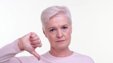 Kararlı, yaşlı, beyaz bir kadın başparmağıyla aşağıya doğru işaret ediyor. Onaylamadığını ve ciddiyetini dile getiriyor. Onun sert ifadesi sert duruşu yansıtıyor, net bir muhalefet anını yakalıyor..