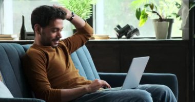 Neşeli Orta Doğulu bir adam kanepeye dizüstü bilgisayarla oturur ve pozitif enerji yayar. Gülüşü ve rahat duruşu iş ve konforu karıştırıp uyumlu bir çalışma ortamı yaratmanın rahatlığını gösteriyor..