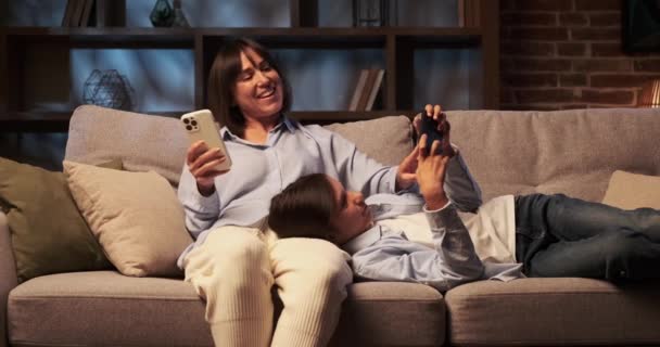 息子と母親はソファーで静かな瞬間を共有し それぞれが携帯電話に魅了されました スクリーンの柔らかい光が顔を照らし 焦点と瞬間の共有された静けさを強調します — ストック動画