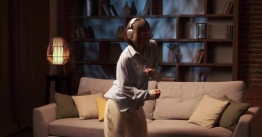 Neşeli Kafkasyalı kız, müzik ritmine uyum sağlayan kulaklıklar takarak oturma odasında özgürce dans ediyor. Yüzü mutlulukla ışıldıyor ve vücudu zarifçe ritme göre hareket ediyor..