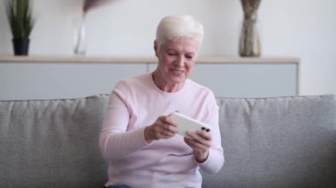 Yaşlı beyaz kadın rahat bir noktada oturuyor, yüzü telefon ekranının yumuşak parıltısıyla aydınlanıyor. Gözler odaklanmış, ve ince bir gülümseme dudakları şereflendirir telefonda bir oyuna daldığında..