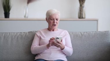 Neşeli beyaz kadın koltukta oturuyor, telefon elinde, yüzü içten ve samimi bir gülümsemeyle aydınlanıyor. Gözleri mutluluktan parıldıyor ve gülümsüyor..