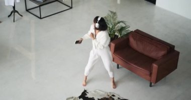 Kulaklık takan neşeli beyaz kadın, oturma odasının sınırlarında dans ederek mutluluğunu ifade ediyor. Hareketleri ve bulaşıcı enerjisi odayı aydınlatıyor..