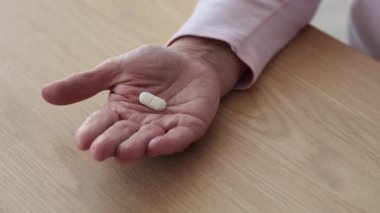 Üst düzey beyaz bir kadının eli nazikçe masaya uzanmış, birkaç hap içiyordu. Yumuşak ışık el çizgilerindeki bilgeliği vurgular, taşıdığı yılların deneyimini vurgular..