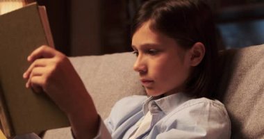 Çocuk, elinde tuttuğu bir kitabın sayfaları karşısında büyülenmiş bir şekilde oturma odasında oturuyor. Gözleri sözlere sabittir ve yüzü hayal dünyasına dalmanın neşesini yansıtır..