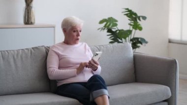 Olgun kadın koltukta oturur, telefonda rahatsız edici haberleri emerken kaşları hoşnutsuzluktan çatılır. Yüzündeki gerilim aşikardır ve gözler endişe ve tatminsizliğin karışımını yansıtır..