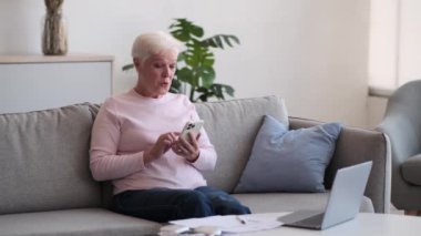 Tatmin olmuş yaşlı kadın koltukta oturuyor, telefonu kullanırken yüzü sevinçle parlıyor. Gözlerindeki onay pırıltısıyla, kendine güvenen bir pozitiflik jesti ile parmağını kaldırır..