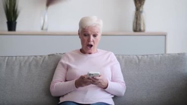 Şaşkın yaşlı kadın koltukta oturuyor, telefon elinde, gözleri şaşkınlık içinde. İfade, gerçek bir şaşkınlık anını yakalar ve duruş, merak ve merak karışımını taşır..