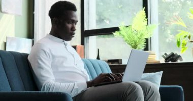 Ciddi bir siyahi adam işine odaklanmış, koltukta otururken dizüstü bilgisayarda yazı yazıyor. Konsantrasyon ve verimlilik sahnesinde görevine olan bağlılığı çok açık..