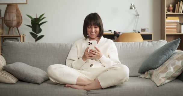 陽気な女性はリビングルームのソファーに座り 電話でテキストを送るように微笑んでいます 彼女の楽しい表情は デジタルコミュニケーションの快適さを捉えています — ストック動画