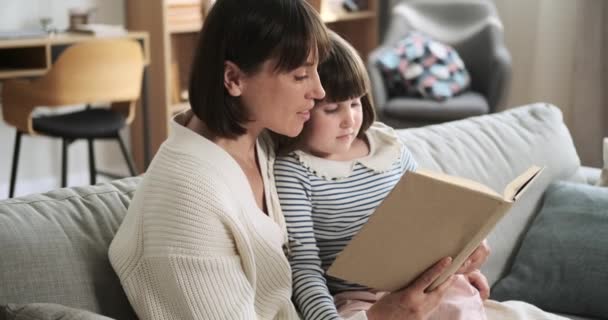 母と娘は 本に没頭したリビングルームのソファーで一瞬を共有します 読書に対する彼らの共通の愛は 家族内の絆と知的探求の心温まるシーンを生み出します — ストック動画