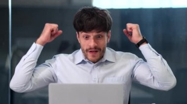 Zafer anında, başarılı bir iş adamı dizüstü bilgisayar ekranına bakarken ışıltılı bir gülümsemeyle ışıldar. Gözleri memnuniyeti ve başarıyı yansıtıyor..