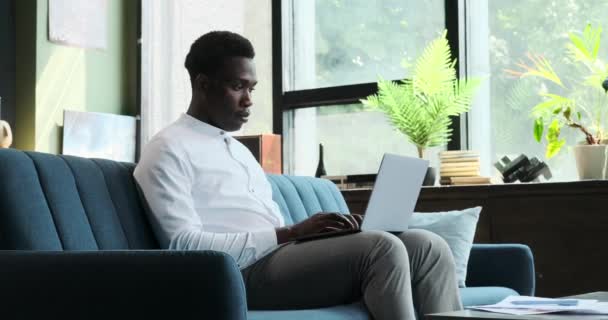 リビングルームの穏やかな雰囲気の中で 深刻なアフリカ系アメリカ人がソファに座り ラップトップのタイプとして仕事に魅了されています 彼の絶え間ない焦点は生産性の雰囲気を生み出します — ストック動画