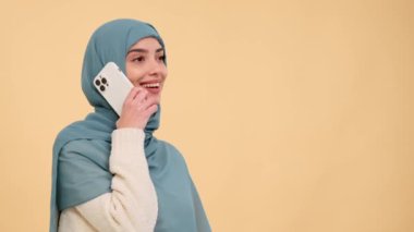 Bir tebessüm ile, tesettürlü bir Arap kadın bej bir arka plan üzerine bir telefon görüşmesi yapar, bu iç açıcı anda pozitiflik ve bağlantı yayar keyifli bir ortamda yakalanır..