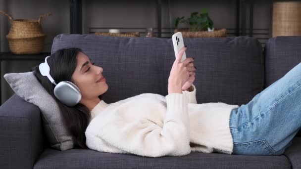 Γυναίκα Που Φοράει Ακουστικά Κοιτάζει Τηλέφωνο Που Βρίσκεται Στον Καναπέ — Αρχείο Βίντεο