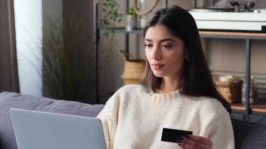 Beyaz bir kadın satın almak için dizüstü bilgisayar ve kredi kartı kullanıyor. Bu resim teknoloji ve ticaretin bütünleşmesini gösteriyor. Oturma odasını güvenli çevrimiçi işlemler için bir alan olarak gösteriyor..