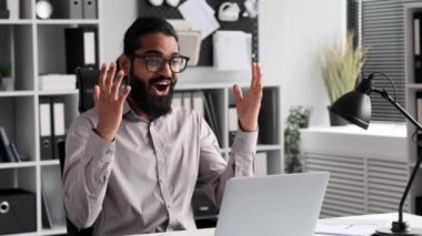 Sevinçli iş adamı, iş kıyafetleri içinde sıralanmış, ofis masasında elde ettiği başarıları bir dizüstü bilgisayarla sevinçle kutluyor. Etkileyici jestler ve keyifli ifadeler bir başarı hissi taşır..