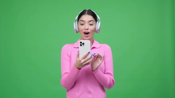 ヘッドフォンを着て携帯電話を持っている白人女性は 良いニュースに驚き 肯定的な感情と緑の背景に本物の喜びで満たされた視覚的に魅力的なシーンを作成します — ストック動画