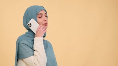 Neşeli bir Arap kadını telefonda sıcak bir bej arka plana karşı sohbet eder, mutluluk saçar ve keyifli bir ortamda canlandırıcı bir an yaratır..