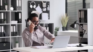 Hintli işadamı ofiste dizüstü bilgisayarını etkin bir şekilde kullanırken bir telefon görüşmesi yapıyor. Çoklu görev gösterisi profesyonellik ve pozitifliğin uyumlu bir karışımı.